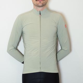 A white man in a sage green long sleeve cycling jersey and black cycling shorts stands against a white wall