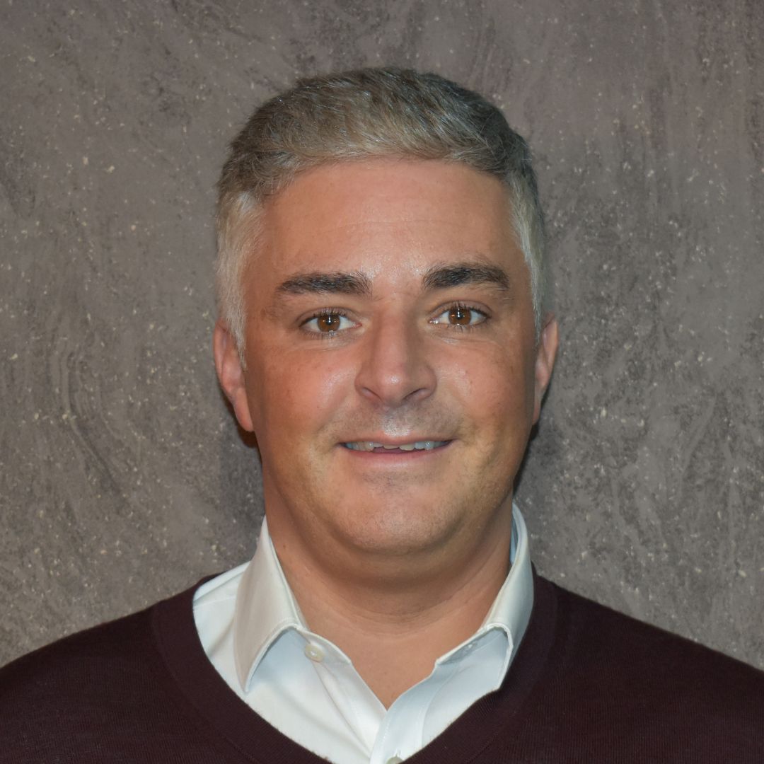 A man with grey hair wearing a white collared shirt and a black jumper smiles at the camera