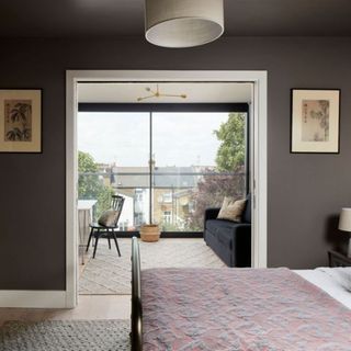 View from bedroom with double bed and dark grey walls through open pocket doors to room with grey sofa, chair and table and full height doors to exterior