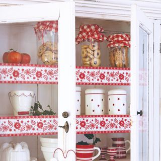 A white Christmas cupboard with glass inserts decorated for Christmas with glass jars of cookies set on the top shelf