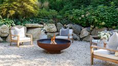 Four outdoor wooden chairs with deep white cushions and throw pillows, spaced out around a low-profile round metal fire pit. The floor is pebbled, there is a stacked rock border and everygreen shrubs in the background