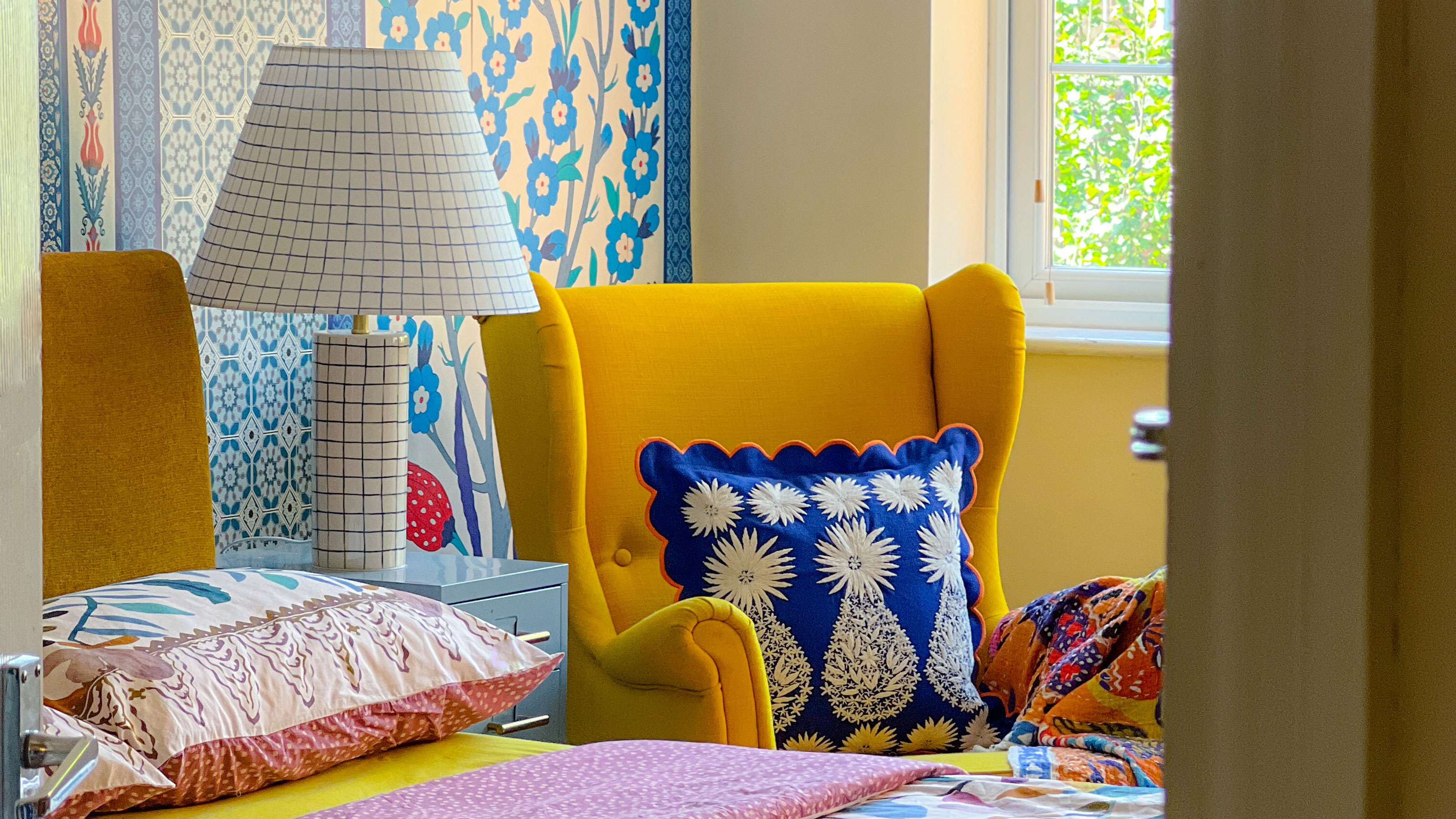 Bedroom with floral wall behind bed, floral duvet cover, checked bedside light and yellow chair with blue cushion