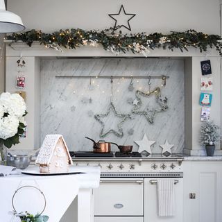 White oven range in the chimney hearth decorated with lights, star shapes and foliage, white table with gingerbread house on the table in the foreground