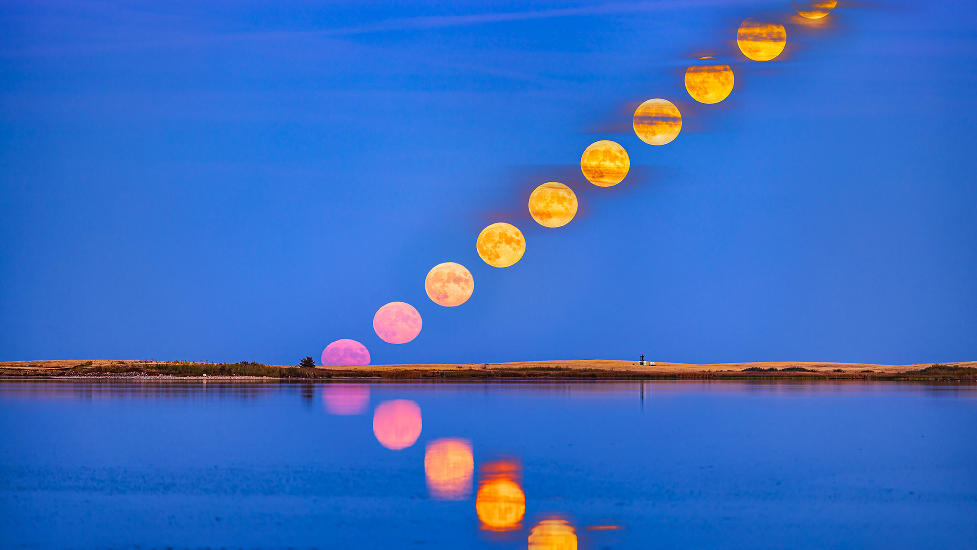 The rising of the full Hunter's Moon of October 9, 2022, with the moon reflected in the calm waters this night at Crawling Lake Reservoir in southern Alberta, Canada. The moon appeared very pink as it rose into the clear sky near the horizo, then turned more yellow as it rose into scattered bands of cloud.