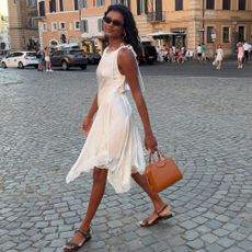 Simone Ashley wears a white lace-trim dress with black sandals and a tan bag as she walks through the streets in Italy.
