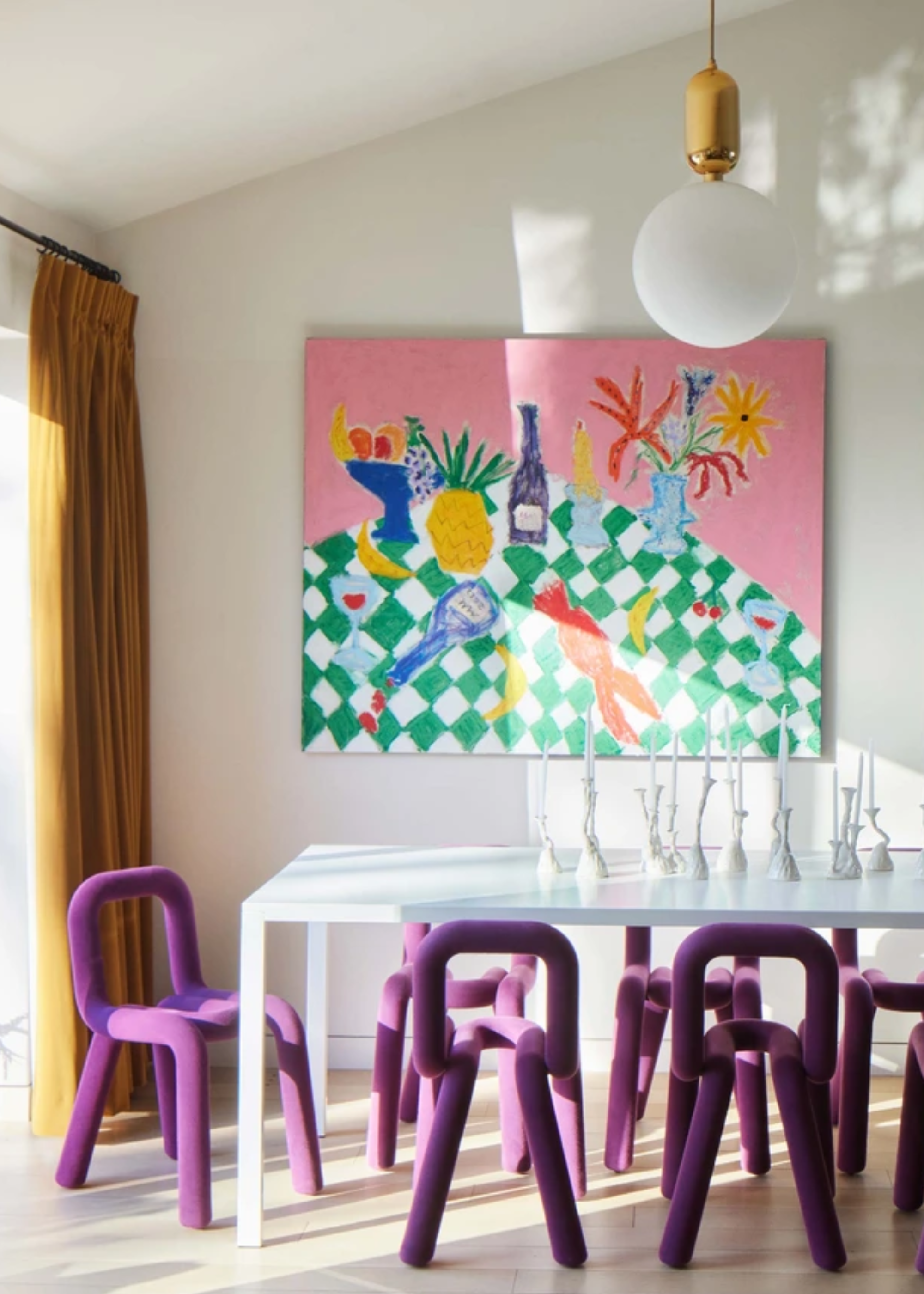 A bright dining room with mustard curtains, a hanging pendant, a light blue dining table with tubular fuzzy purple chairs, a collection of white vases and a painting of a checkerboard table with fruits, a vase, lobster and wine on the wall