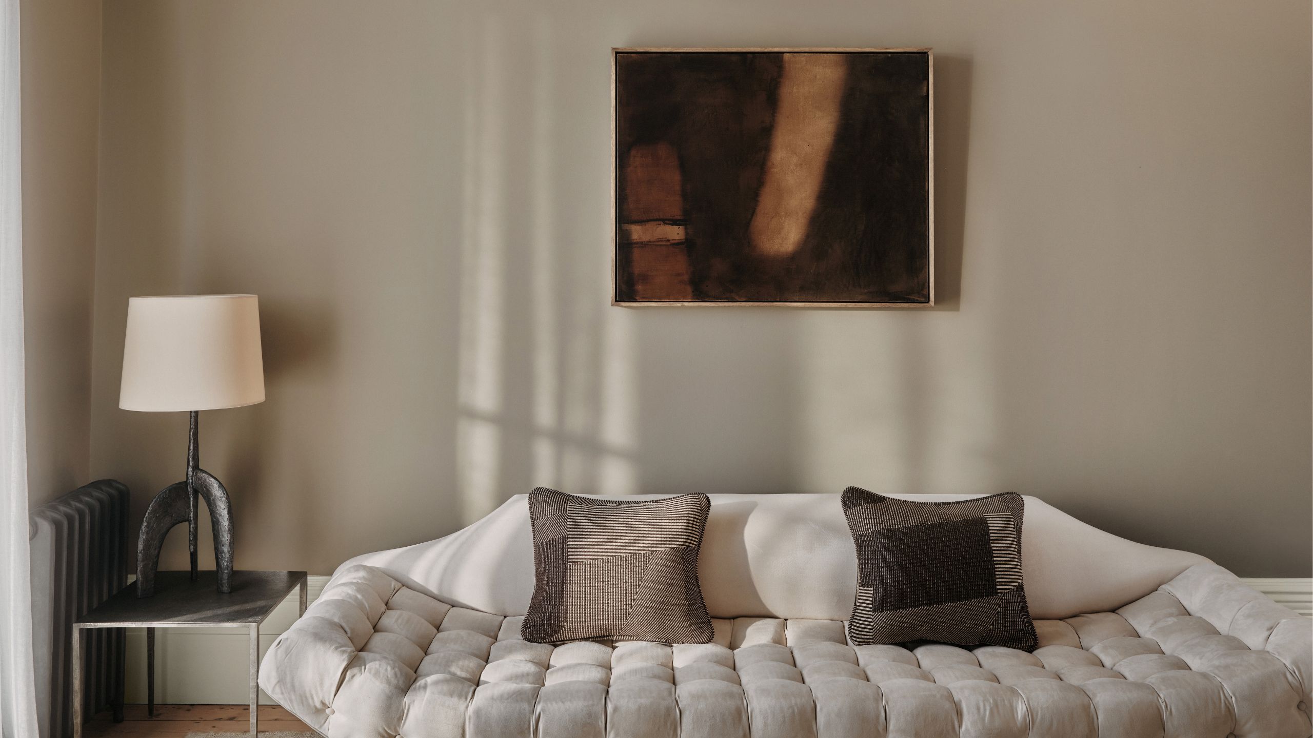 Living room painted in Mouse's Back by Farrow &amp; Ball, a cream sofa with a quilted base, a side table with a metal hammered lamp on top, and an abstract dark brown painting.