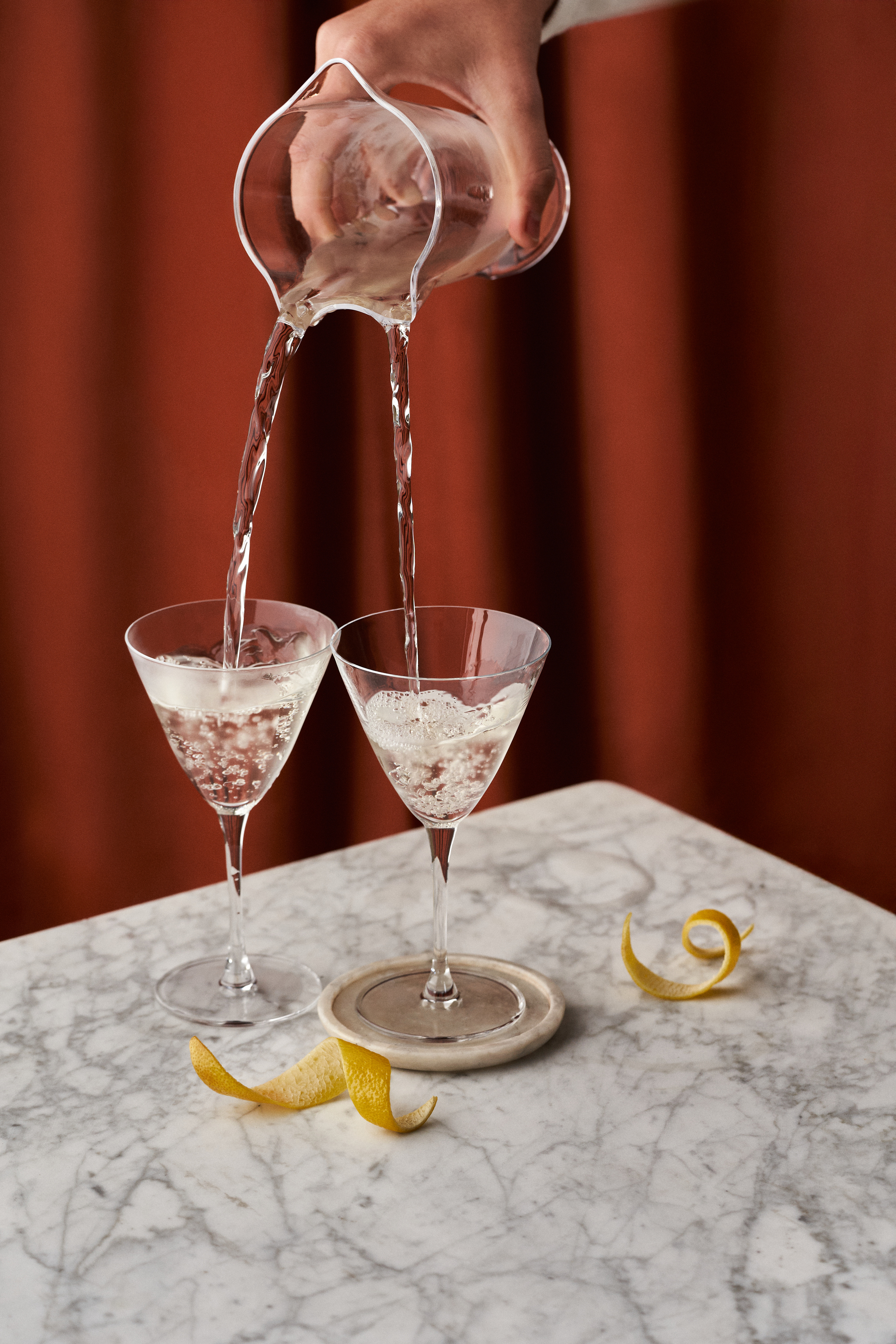 Image of a hand pouring martinis into two martini glasses from a single pitcher. The glasses are on a marble countertop in front of a red background.