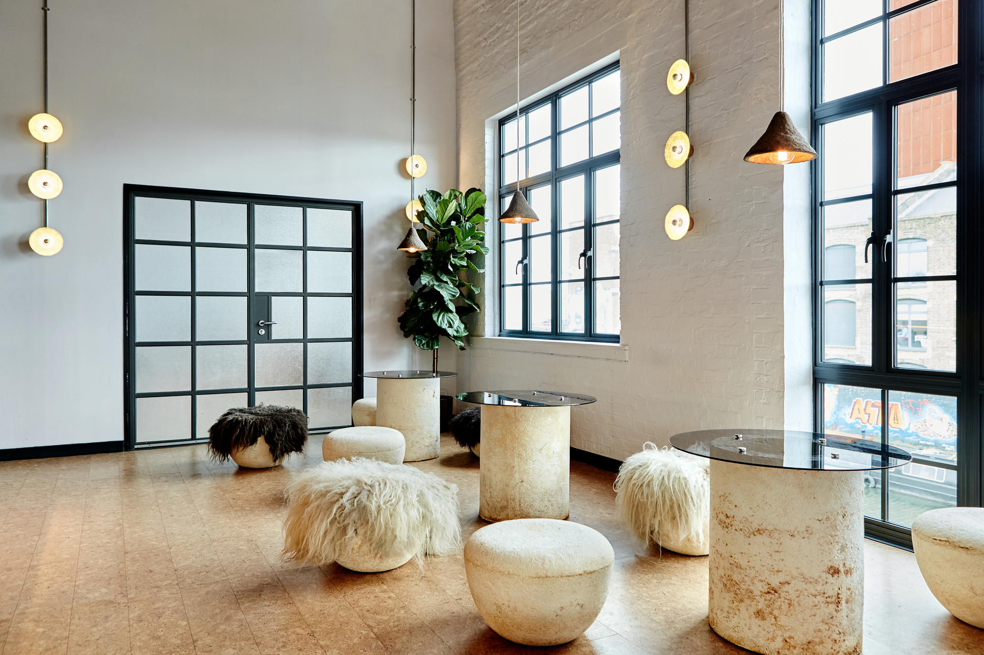 a cocktail bar with travertine tables with glass tops, cork flooring, stone stools with sheepskin, large steel-framed windows, pendant lights, and a potted fiddleleaf in the corner