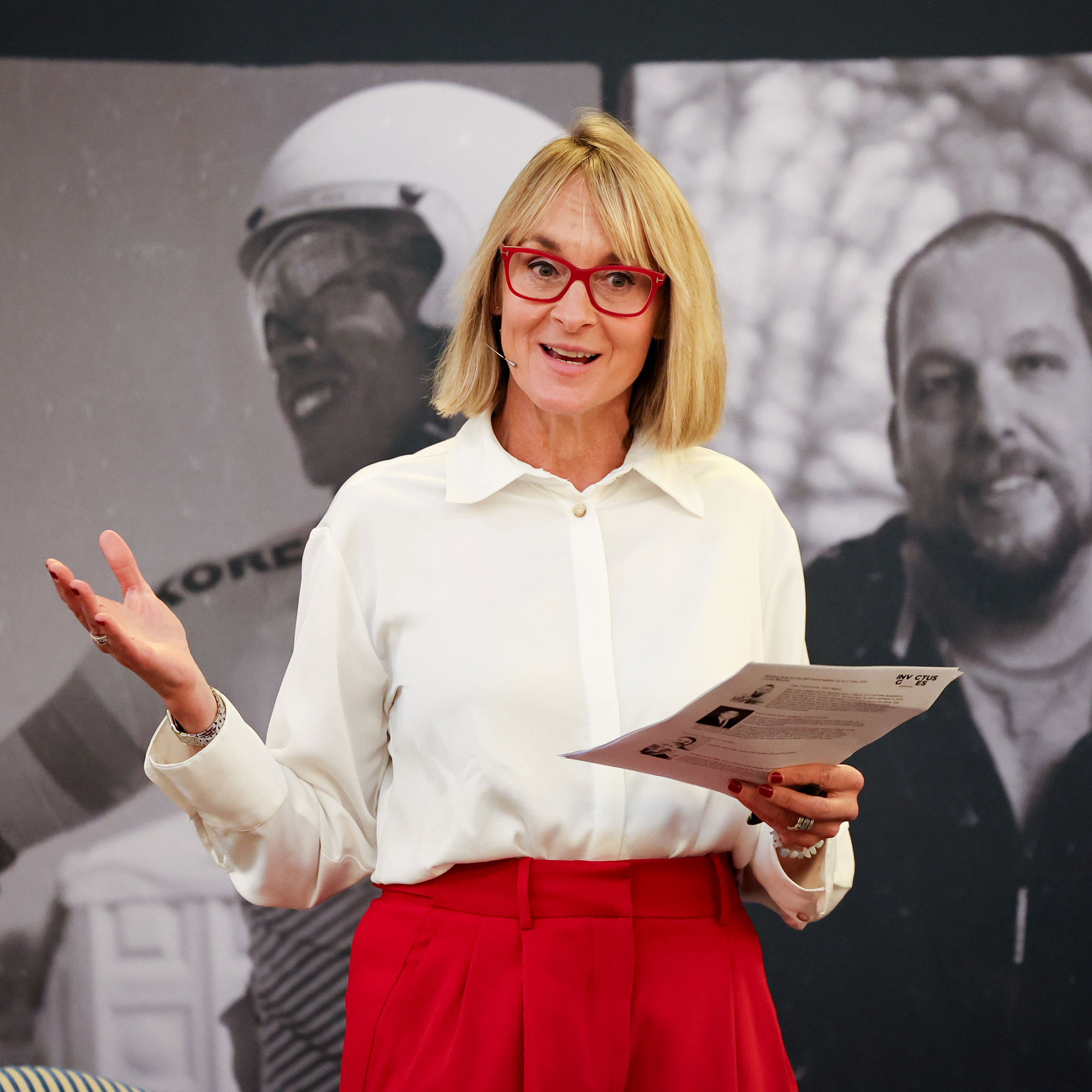 Louise Minchin in a white shirt and red trousers, in front of a black and white background photo of a man and woman