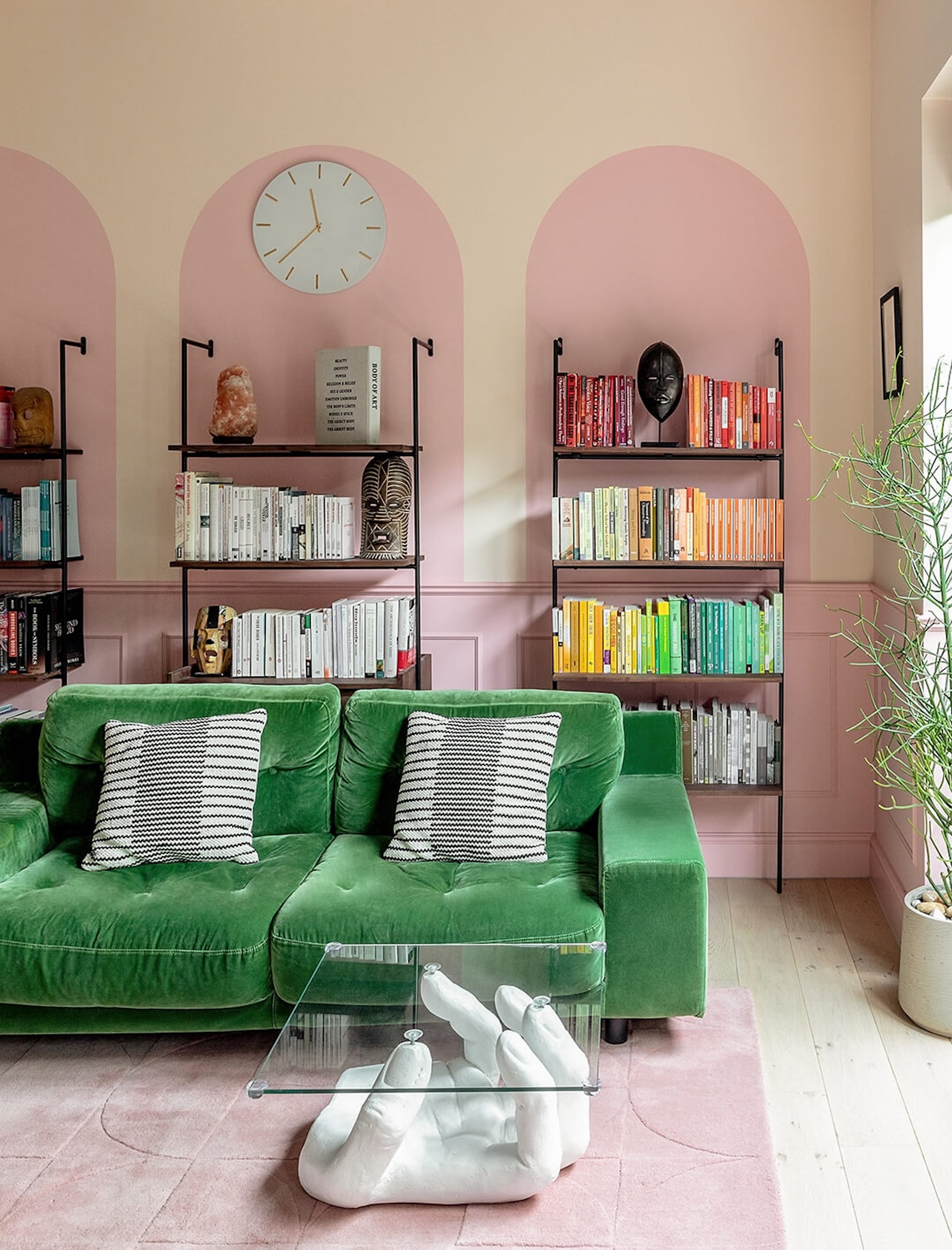 A pink toned living room with curved details on the far wall, and black framed book shelves as well as a green sofa in front and a modern coffee table.