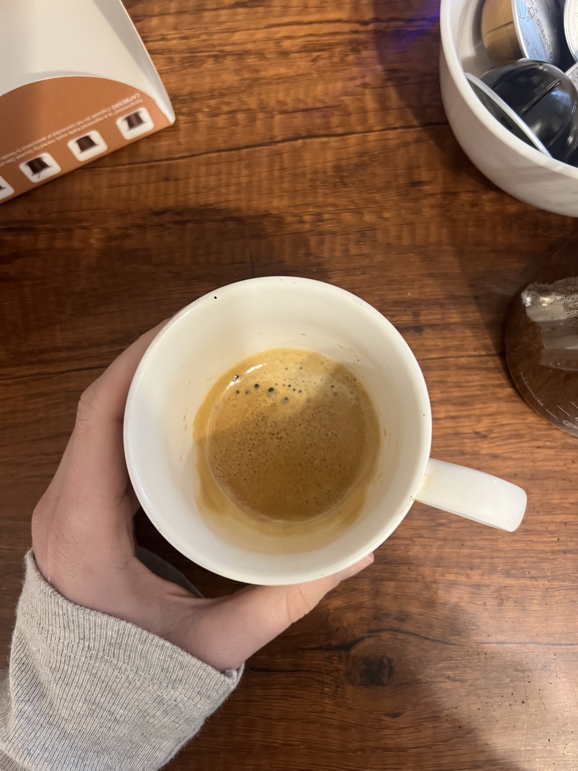 image of a hand wearing a long-sleeved gray shirt holding a coffee mug filled with an espresso shot over a wooden countertop. There is a bowl of coffee pods also on the counter.