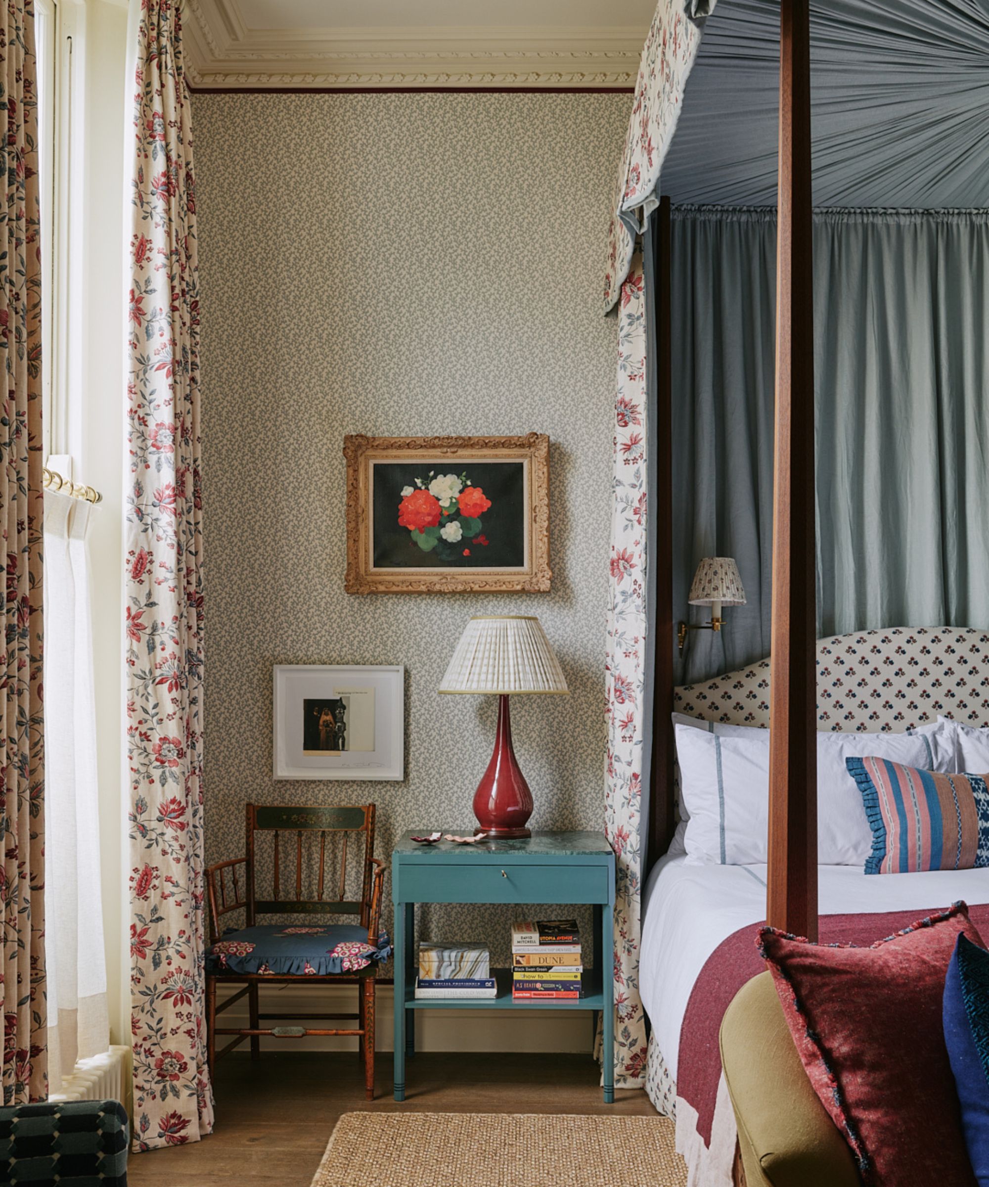Vintage bedroom with four poster bed, floral drapes, blue lacquer nightstand, rattan rug, vintage still life of flowers and antique chair