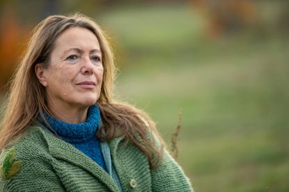 An older woman sits outdoors. It appears to be fall and she is wearing a sweater. Her expression is thoughtful or serious.