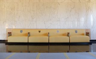 A long, light coloured sofa with four wide seats, armrests and shield style designs on the backrests at Hilversum Town Hall. The space features large marble tiles on the wall and dark tiled flooring