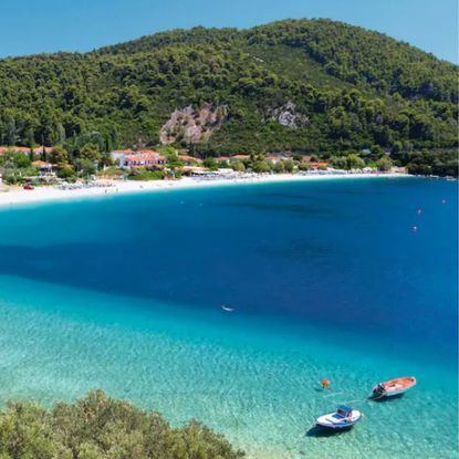 A photo of a beach in Skopelos - blue sea with two small boats, white sand, and a green mountain.