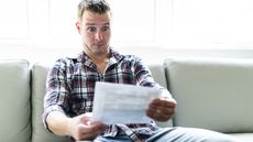 A man looks shocked as he looks at a bill while sitting on the sofa.