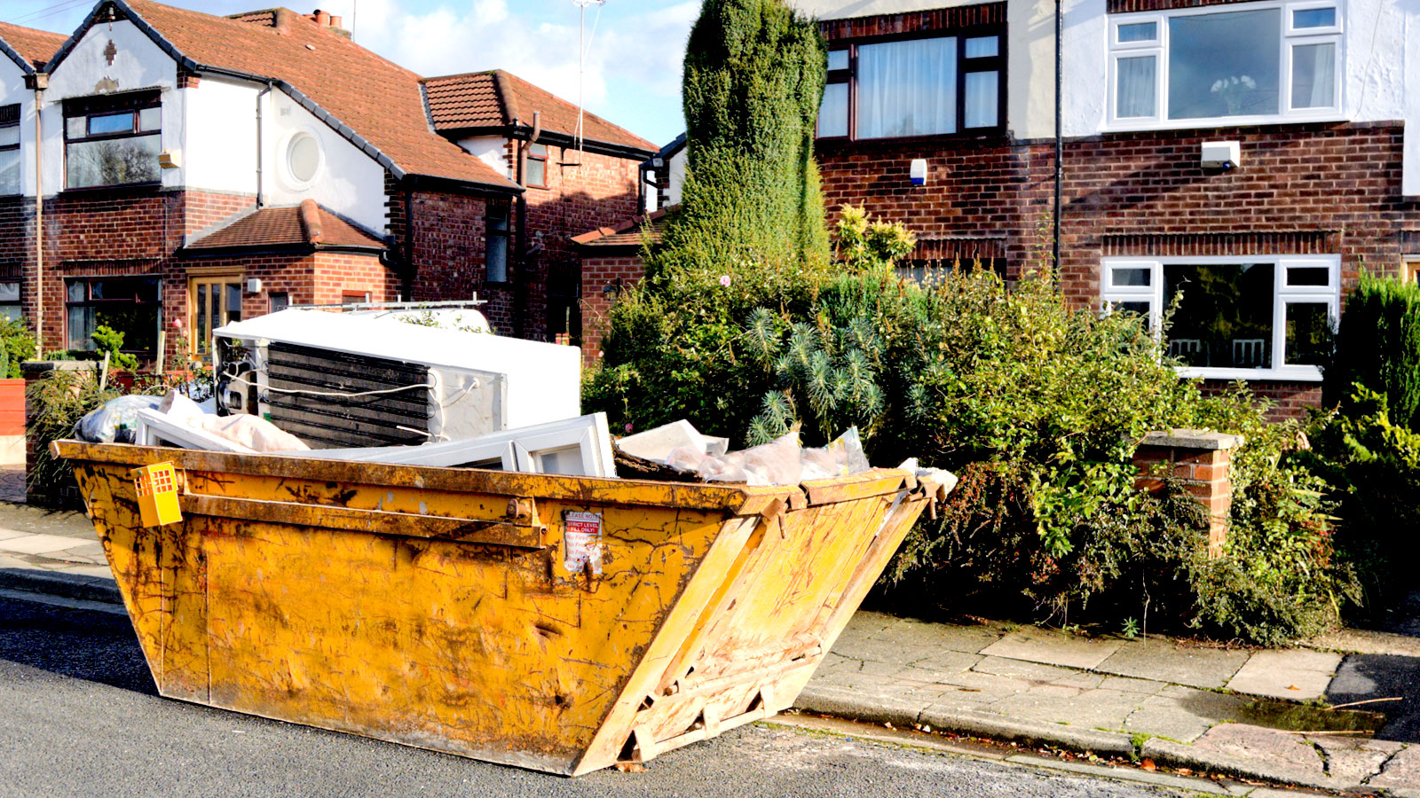 Yellow skip fille with fridge, windows and general waste on road outside a house