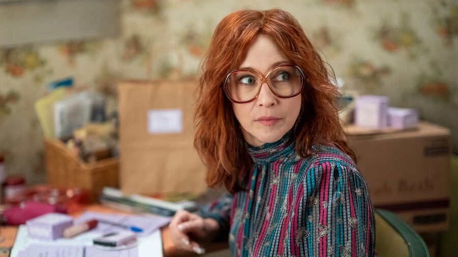 Vera Farmiga sitting at a desk, wearing large glasses and a patterned blouse