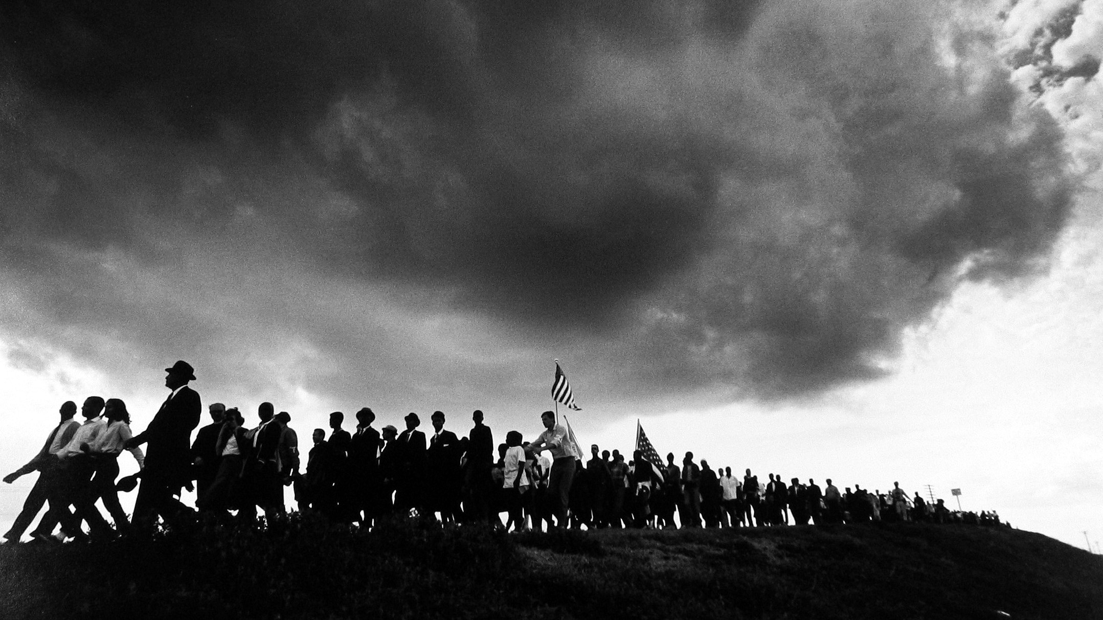 A dense, dark storm cloud looms over a long line of silhouetted marchers carrying American flags as they walk along a ridge during the Selma to Montgomery march.
