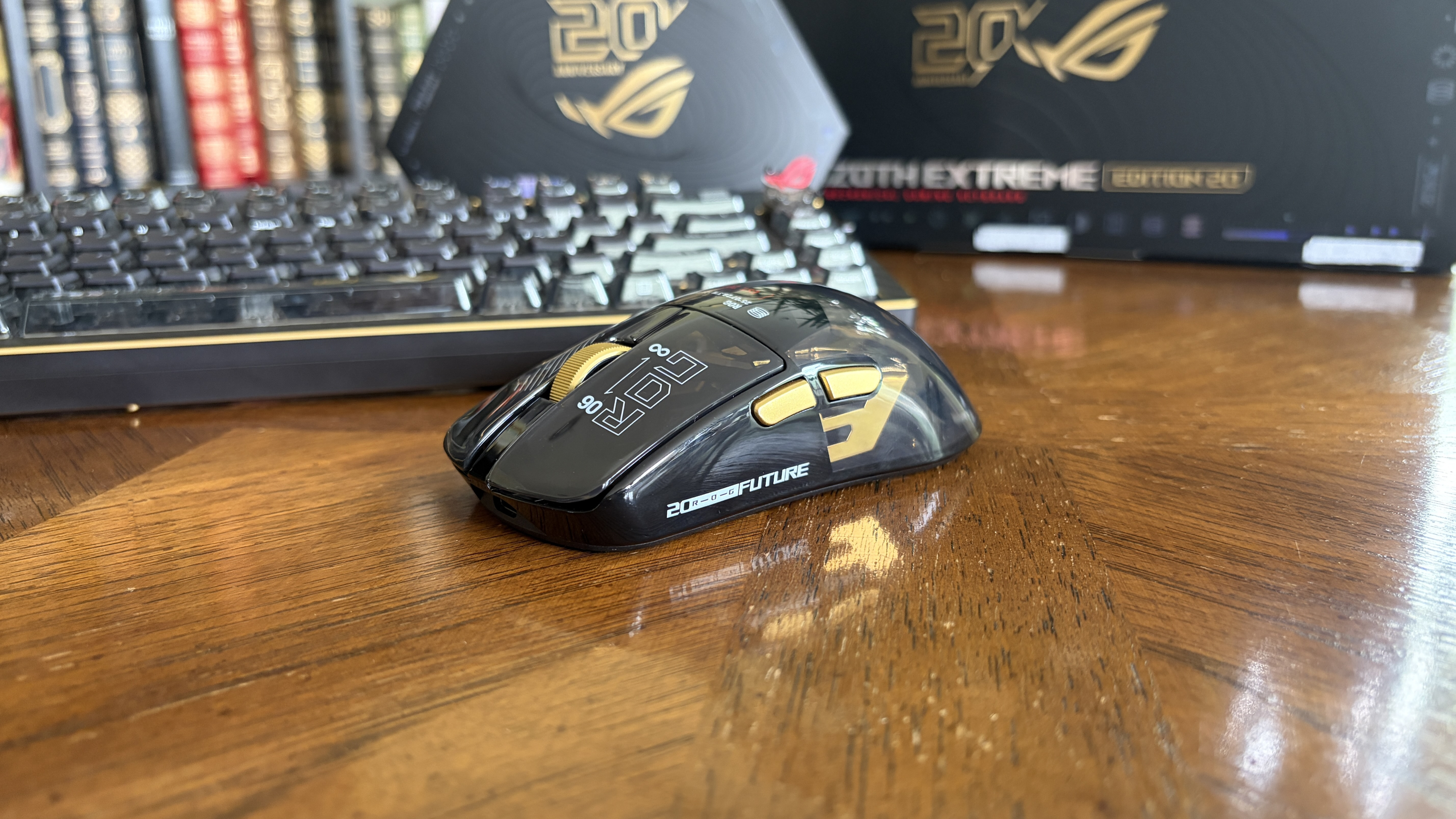 A black and gold ASUS ROG computer mouse on a wooden table, with a keyboard and bookcase in the background.