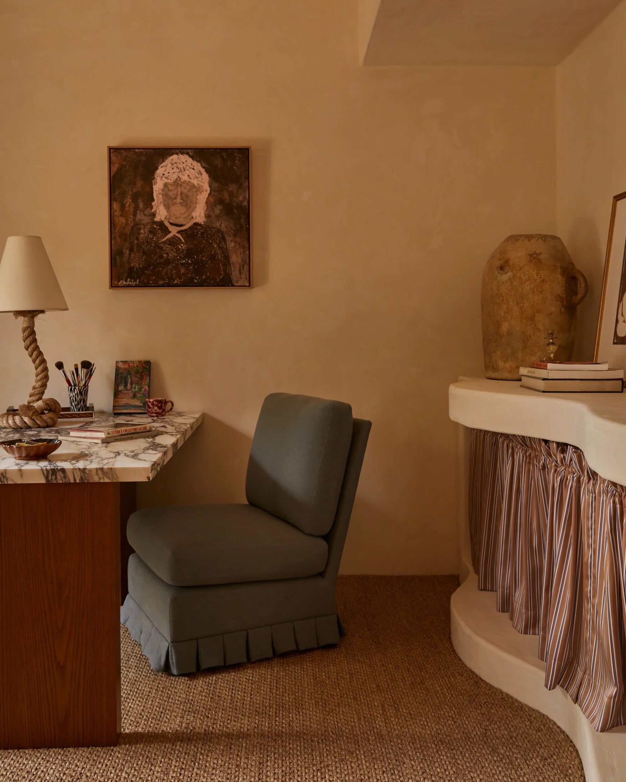 Image of an off-white limewash room with jute rugs, a light blue box pleat chair, a marble and wood desk, a rope table lamp, and a console that has a striped skirt.