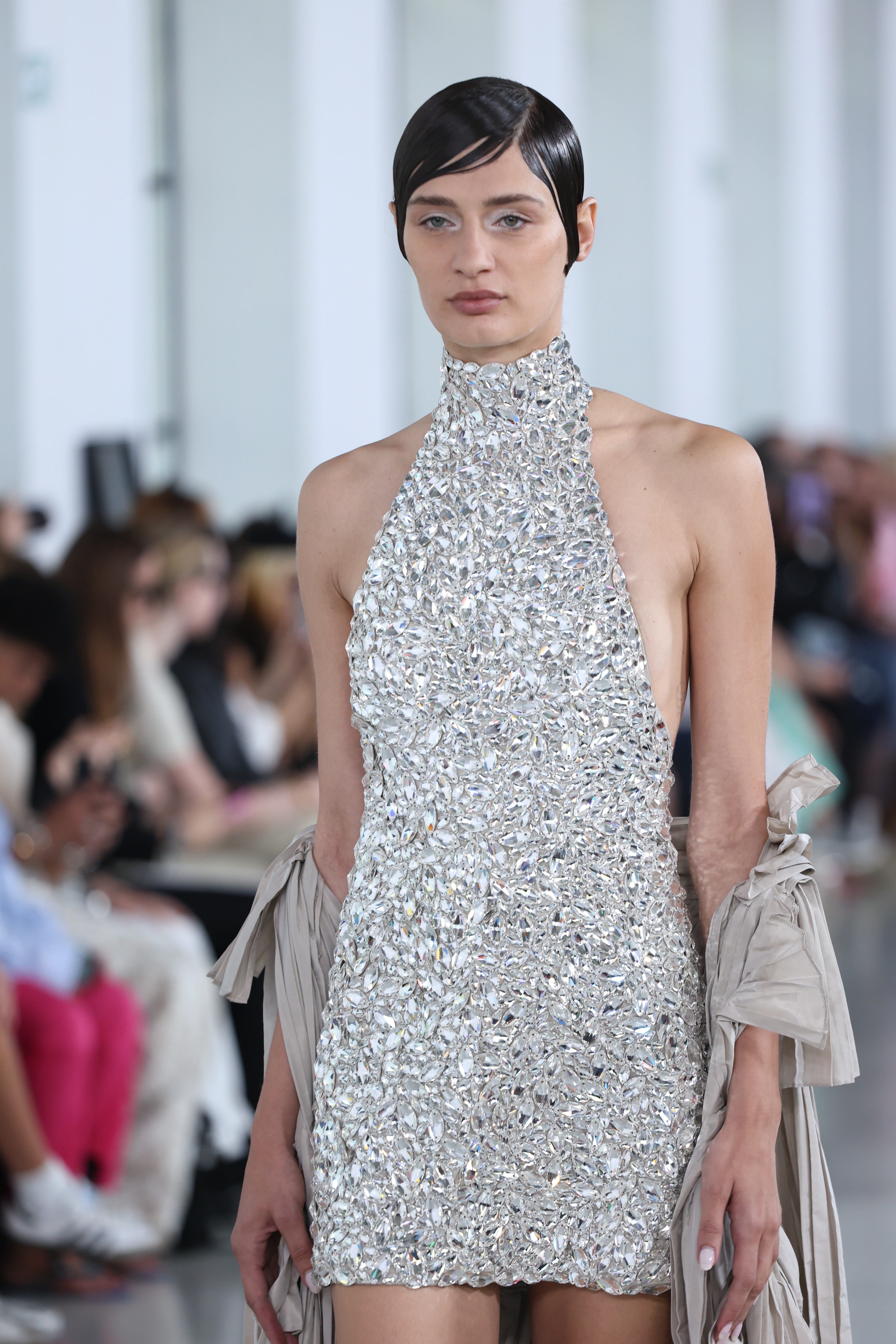 A runway model wearing a silver textured minidress with a high neck