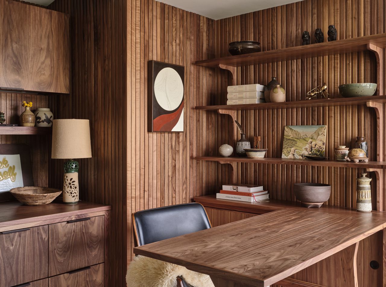 A wooden desk in the corner of a room with panelled walls, a Persian rug, and shelving 