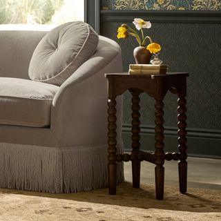 Half of a gray/off-white fringe-trimmed couch with a bobbin, dark-wood side table beside it