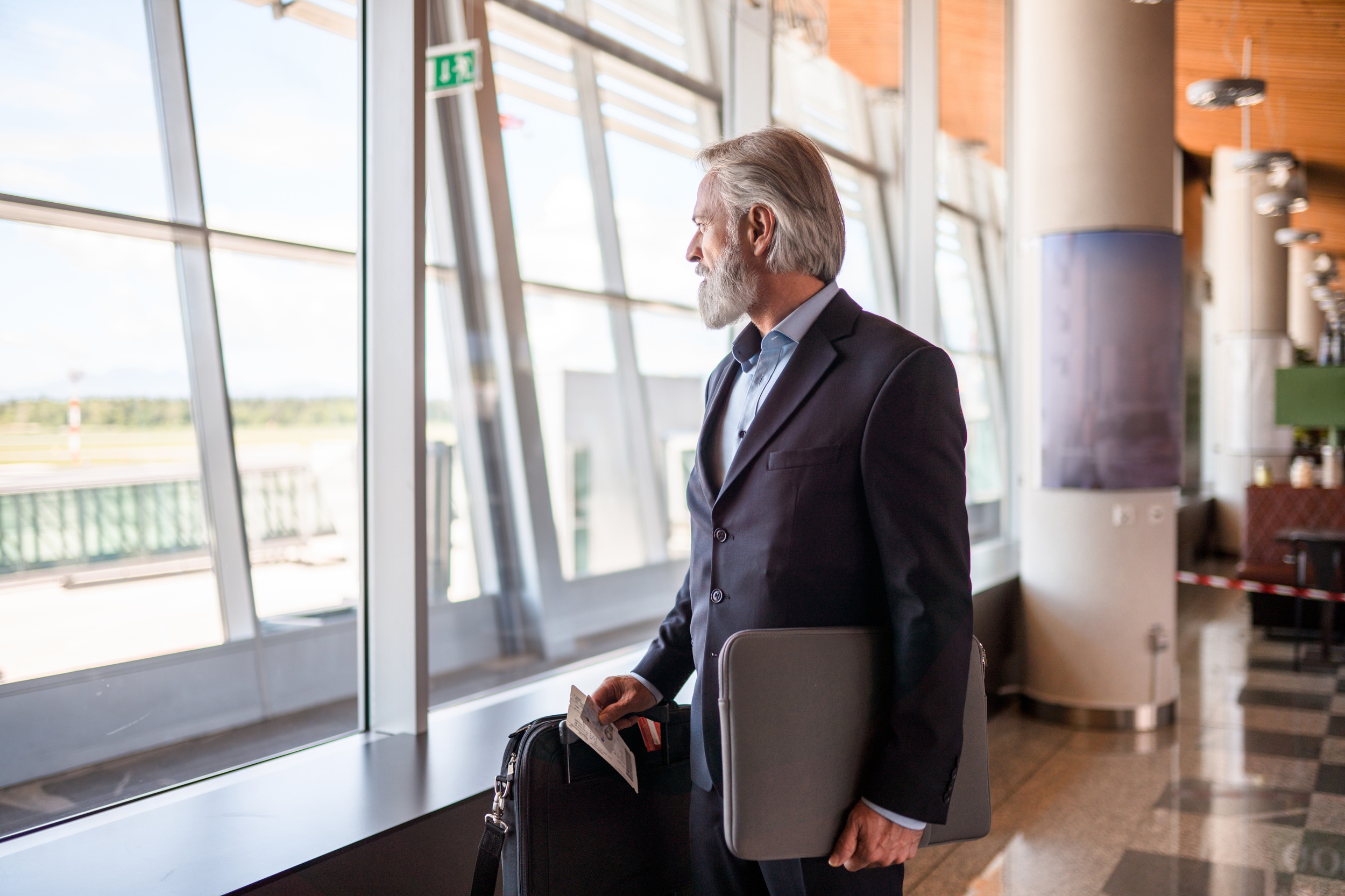 Aged Caucasian business man with beard, wearing a suit and carrying a laptop, looking through a large window at a departure gate. 3/4 length image. Side view.