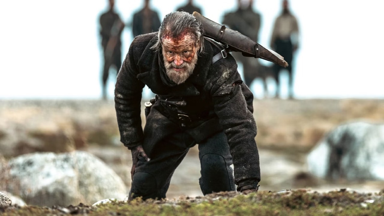 The image shows a bearded man on his knees with blood on his face. Five figures can be seen standing in the background.