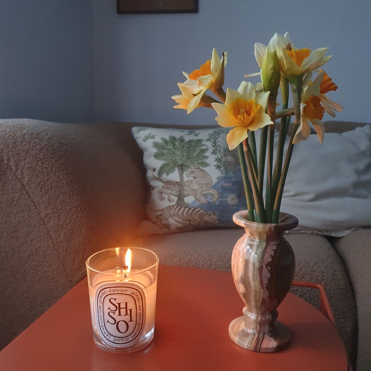 A lit Shiso candle from Diptyque on an orange coffee table next to a bud vase filled with daffodils