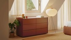 A cinnamon colored dresser with six drawers in a bedroom near a potted plant and a standing rice paper lamp