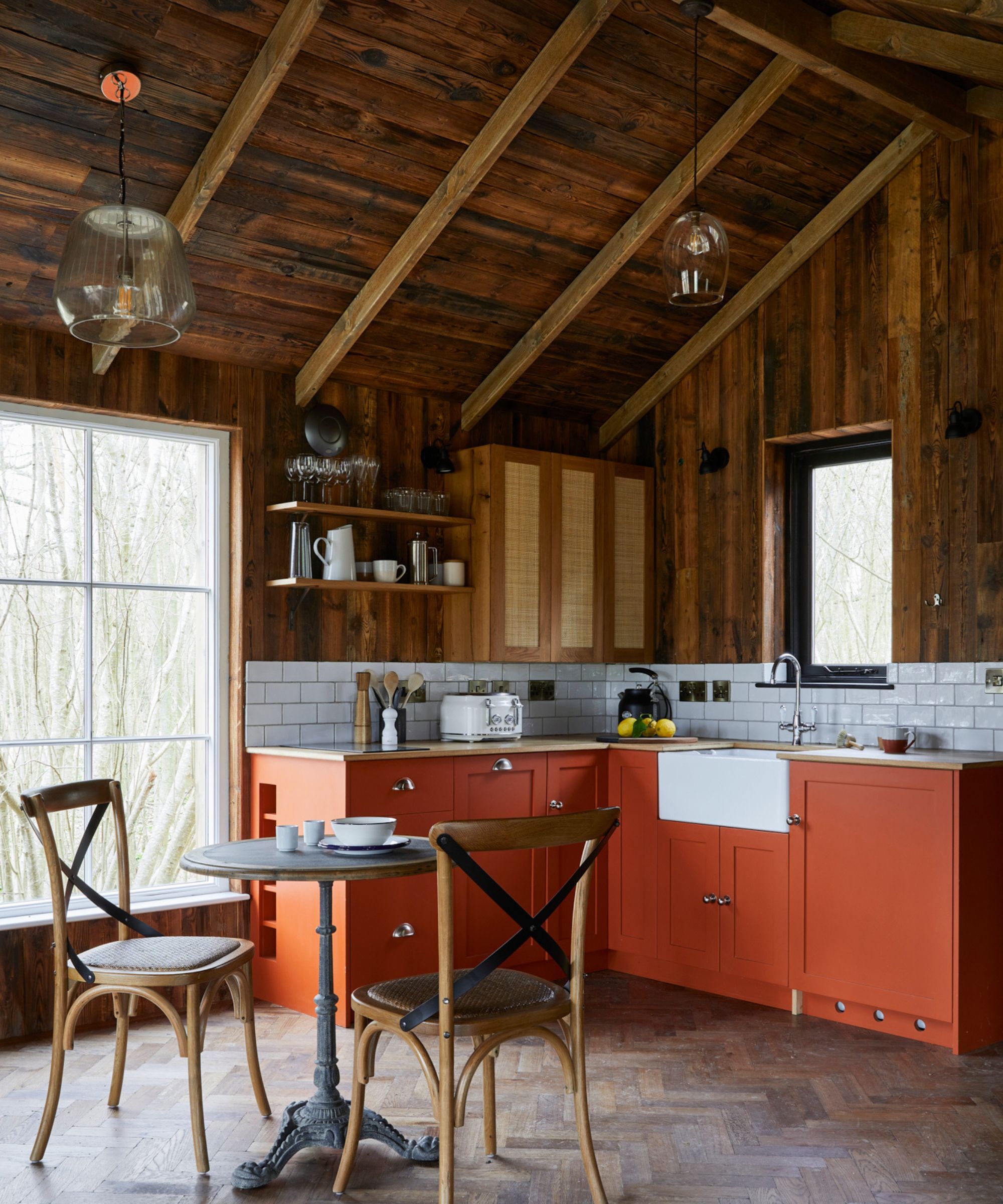 A rustic-meets-modern treehouse kitchen with deep terracotta cabinetry, open shelving, traditional Belfast sink, and rich wood walls. The herringbone floor and iron pedestal table add a heritage feel to the compact space.