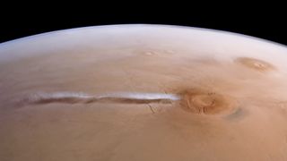 wispy clouds float above a reddish orange planet as seen from space