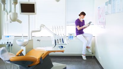 A dentist looks at a tablet in her office, with a dentist chair in the foreground.