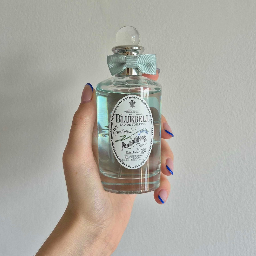 A close-up of Digital Beauty Writer Naomi Jamieson's hand, seen holding a bottle of Penhaligon's Bluebell, which features a pastel blue bow/ in front of a cream wall background