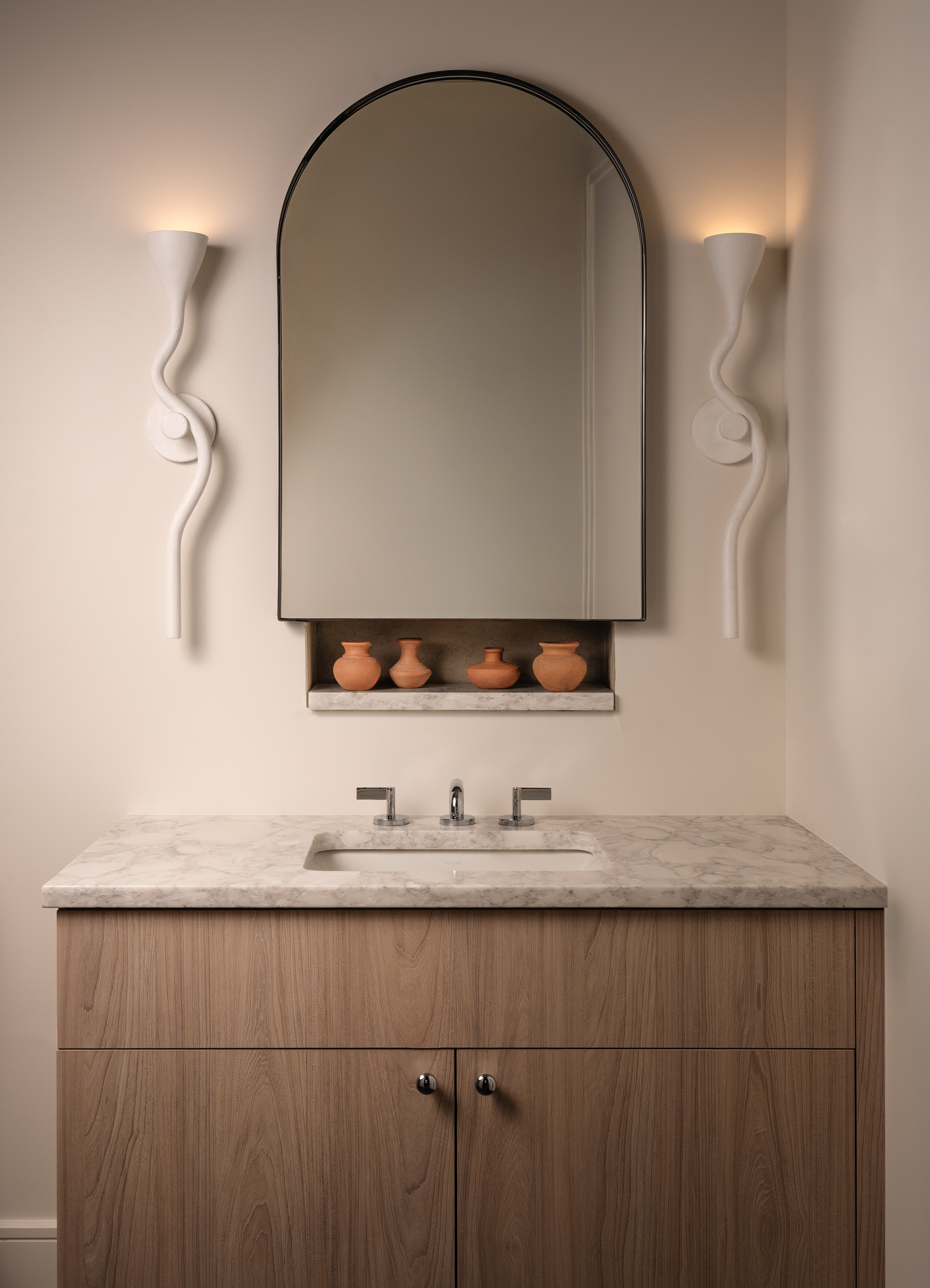 Neutral bathroom vanity area with arched mirror and white lights either side, and a wooden vanity with marble top