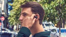 A man with brown hair and a dark shirt wearing JBL Sense Pro over-ear headphones in black in a leafy but urban street