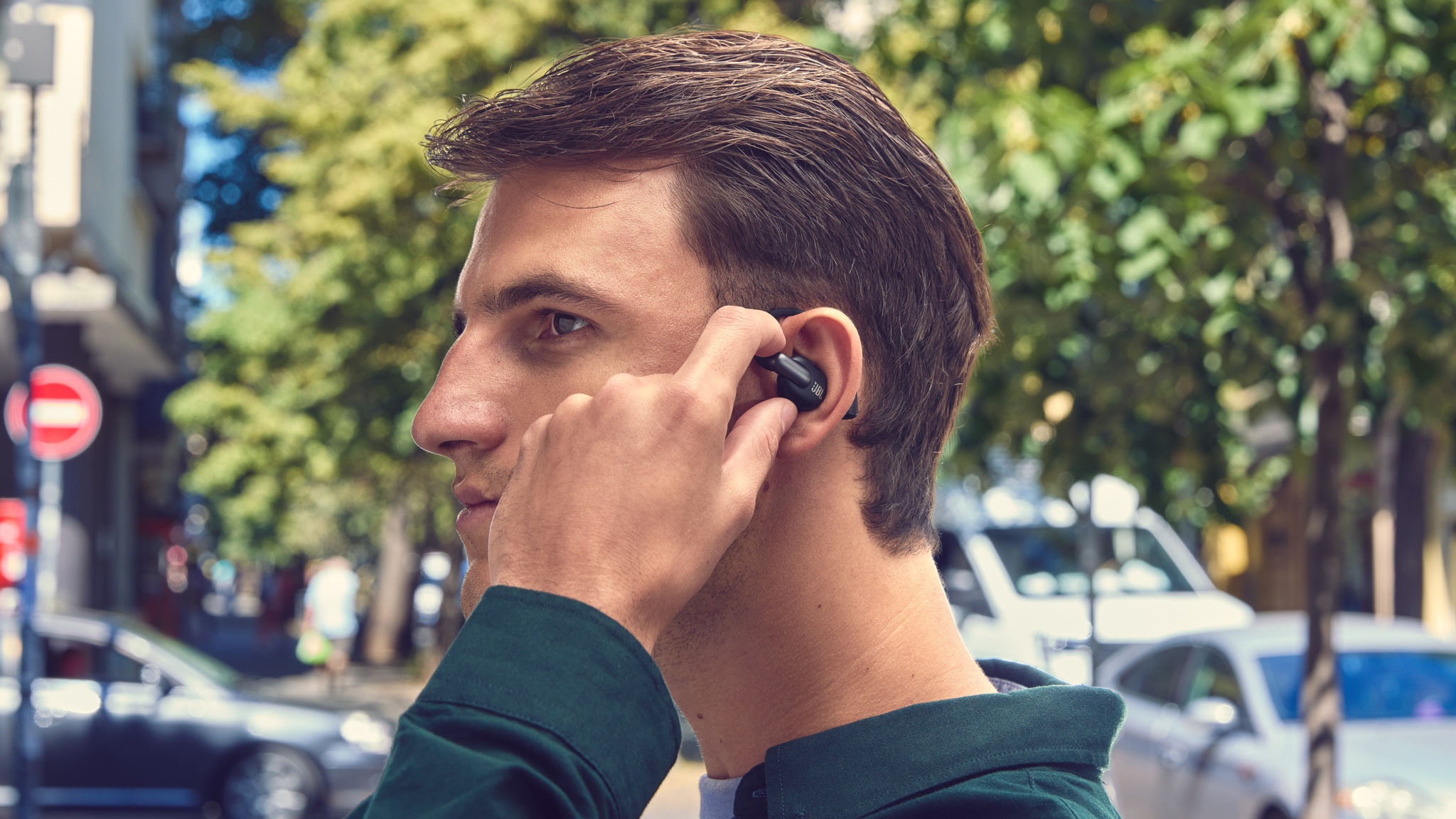 A man with brown hair and a dark shirt wearing JBL Sense Pro over-ear headphones in black in a leafy but urban street