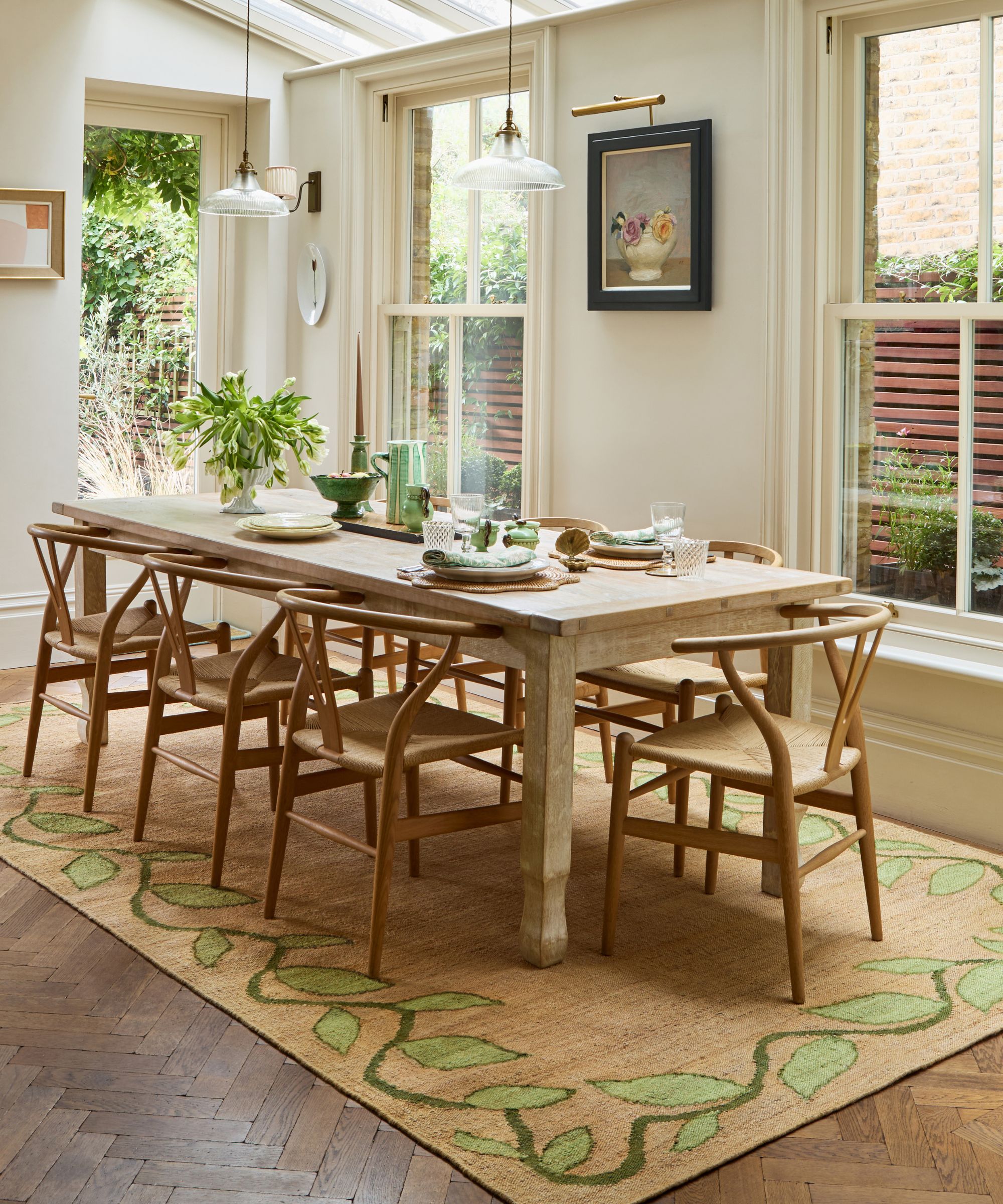 Dining room with skylights and large windows, walls painted cream, low glass pendant lights, a long wooden dining table with wicker dining chairs, and a jute rug with a leaf pattern border underneath