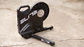 A black Elite Suito Direct Drive Smart Turbo Trainer stands on a concrete floor outdoors