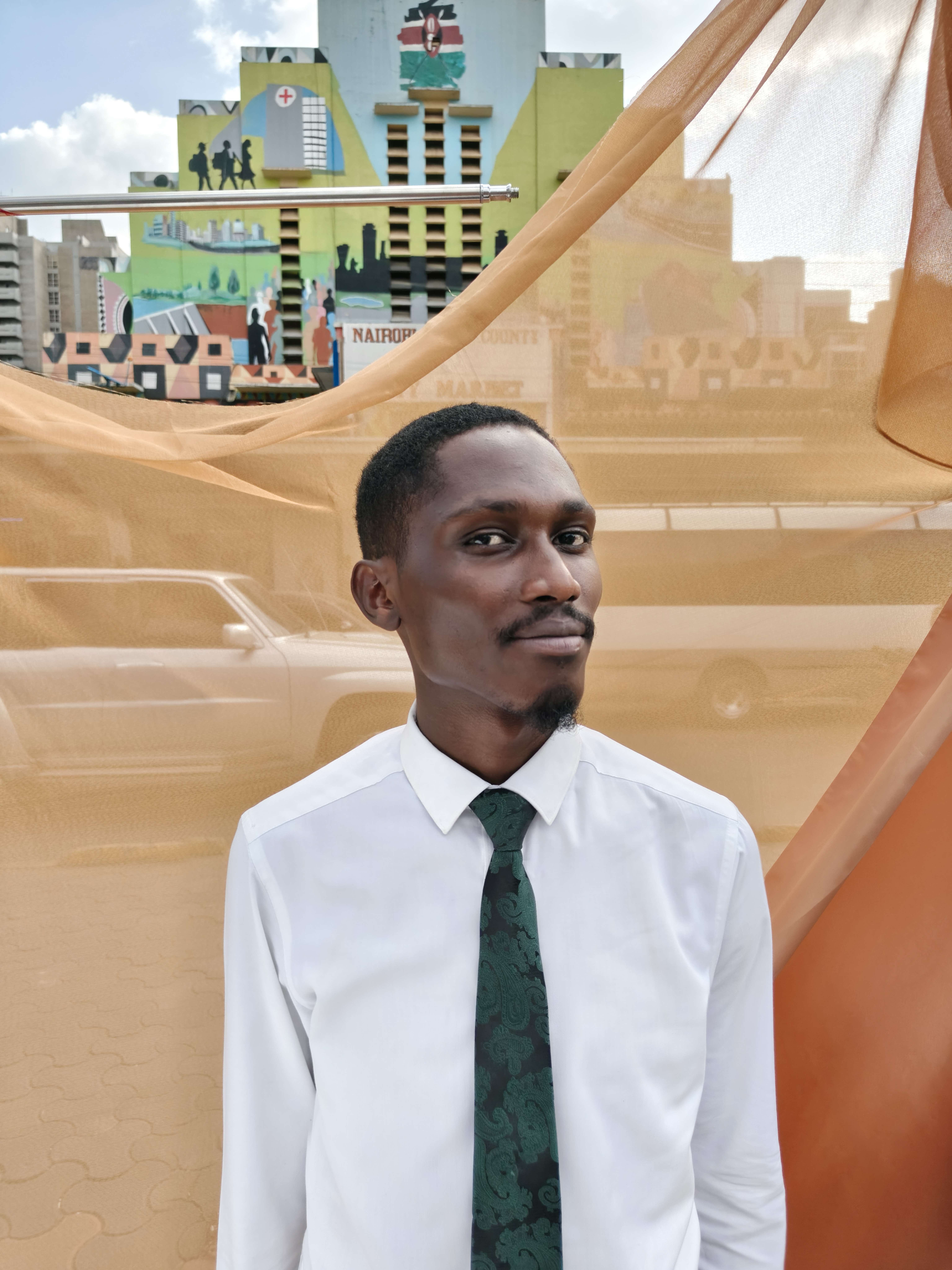 A young man in a white shirt and dark green paisley tie stands in front of sheer orange fabric, with a colourful mural-covered Nairobi County building behind him.