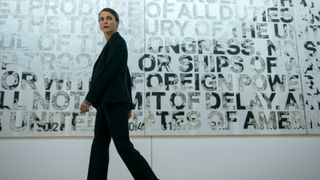 Keri Russell as Kate Wyler wearing a black suit and looking at something off camera as she walks past a poster with numerous words on it.