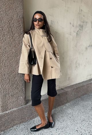 best cropped trench coat for women shown on a woman standing outside against a wall wearing a tan funnel neck trench coat with black capri pants, black ballet flats, and a black frame bag