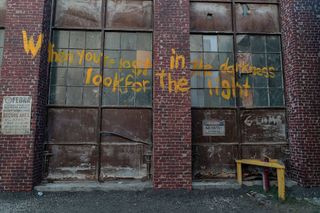 Yellow painted words on the side of a building read "When you're lost in the darkness look for the light" in The Last of Us