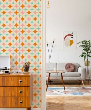 An entryway with orange, pink, and blue star wallpaper and a wooden sideboard, looking onto a living room with a gray couch, a dark brown wooden table, and a geometric rug