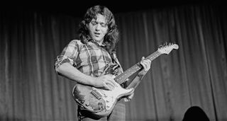 A black and white photo of Rory Gallagher wearing a plaid shirt, playing his '61 Strat