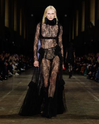 Alex Consani wearing a sheer lace black gown with a ruff while walking at the F/W 25 Alexander McQueen show.