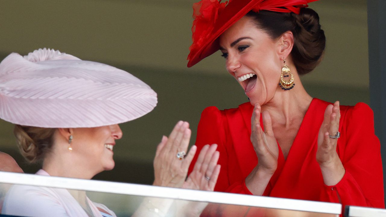 Kate Middleton wearing a red dress and hat clapping and laughing while looking at Duchess Sophie wearing a pink hat and dress at Royal Ascot